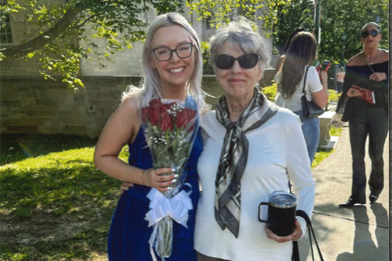 A young woman in a blue dress, holding red roses, beams next to an older woman with sunglasses. 