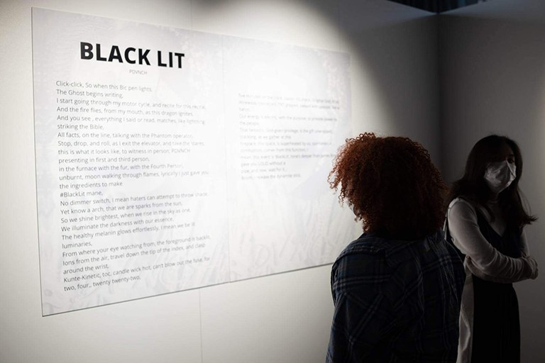 Two people stand in the shadows to read a wall poster with the headline Black Lit illuminated