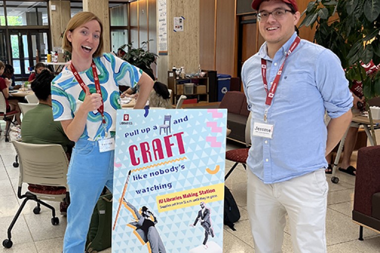 Two people stand happily next to a poster that promotes a crafting activity inside a library lobby