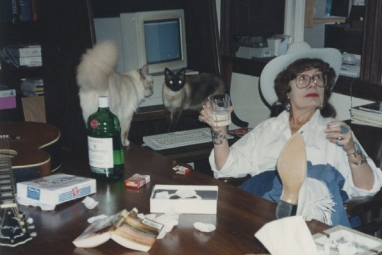 A woman sitting at a writing desk with her feet up, holding a drink, and accompanied by two cats.