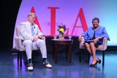Two people are seated in arm chairs for a formal discussion in front of a live audience.  The person on the left wears a suit with a blue tie and the person on the right wears high heels and a blue skirt suit.  In the background large read letters read ALA.