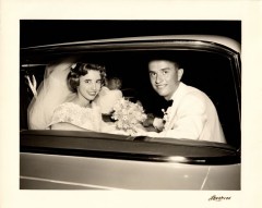 Black and white photo of a bride and her groom looking out the rear window of a car.