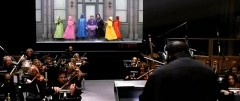 A group of performers stand on a raised stage in a rainbow of dress colors.  In the foreground are musicians playing instruments.