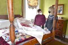 An antique bed in a historic house museum has several quilts on it. Two people stand next to it discussing one of the quilts.