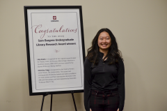 Christina stands next to a large congratulatory sign. She has a big smile. She is dressed in black and is an Asian-American young woman. 