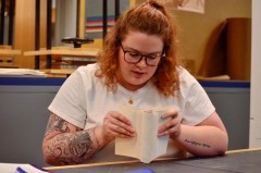 With curly strawberry blond hair, Emily Stanley examines a book, seeing what's needs fixed. She is wearing a white shirt and dark framed glasses.