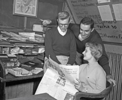 A black and white photo of three young people -- two males and a female-- looking at a newspaper. It is obviously the 1950s due to their hair and clothes. In the background is a messy desk.