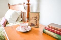 A bedside table in the Wylie House. On it rests a vintage teacup, a sepia-toned photograph, and some books.