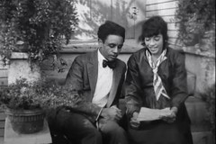In a still from a black-and-white film, two people sit side by side. One person has a paper in her hands. They appear to be conversing.
