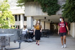 Two students walk out of the patio entrance to the Wells Library. One is wearing tan and black while the other has on an IU shirt. They pass students eating at tables.