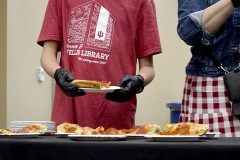 Two gloved hands hold a paper plate with a slice of pizza on it. Several more can be seen on the table in front of the person.