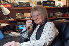 Barbara Yasson sits at her desk with a laptop in front of her, a microphone in her hand, and surrounded by her ham radio equipment.