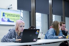Three college students working on laptops with a feminist digital billboard in the background.