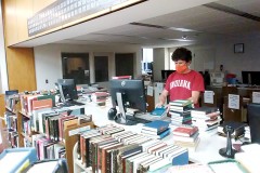 A man wearing a mask is surrounded by books in a library. He uses a computer to scan a barcode.